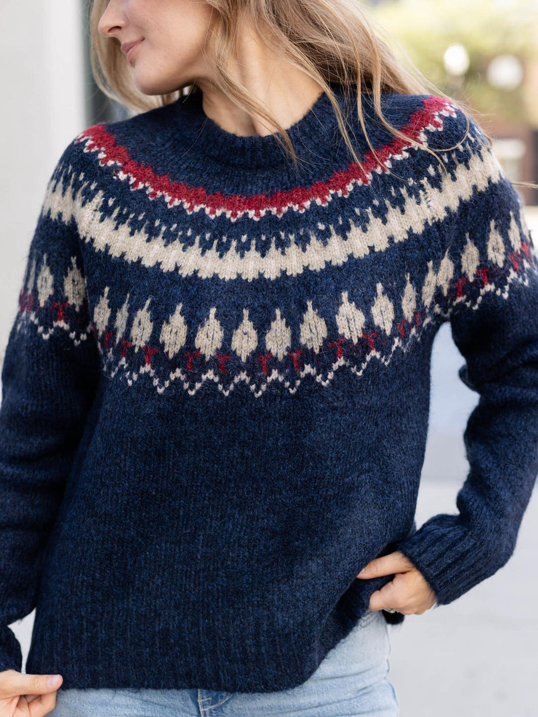A woman wearing a navy blue knit sweater with a traditional Fair Isle pattern in cream and red across the chest and shoulders, paired with light blue jeans.
