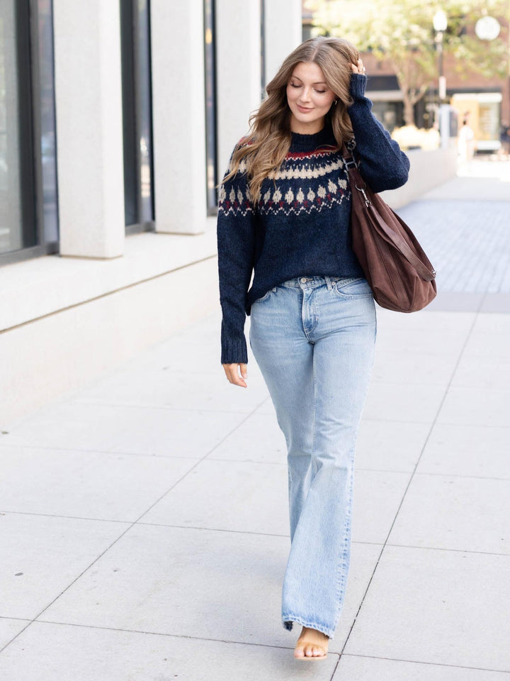 A woman walking barefoot on a city sidewalk, wearing a navy blue knit sweater with a colorful geometric pattern across the chest, light-wash flared jeans, and carrying a brown shoulder bag.
