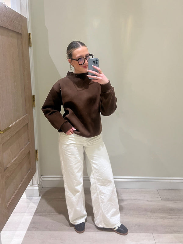 Person wearing a brown sweater and white pants taking a mirror selfie.