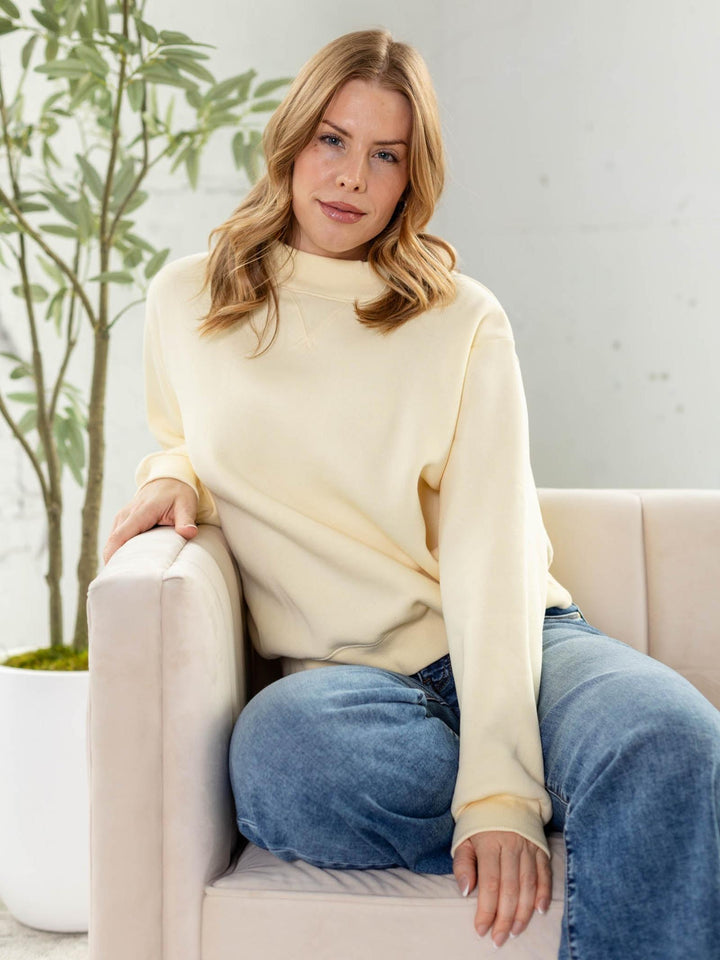 Woman wearing a cream sweater and blue jeans sitting on a beige couch with a plant in the background.
