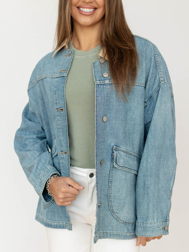 Person wearing a denim jacket over a green shirt and white pants on a plain background
