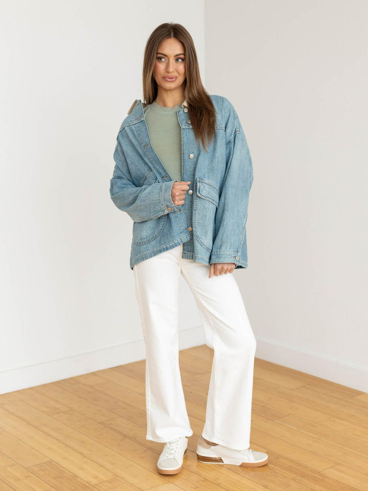 Woman wearing a denim jacket, green top, and white pants in a minimalistic room.
