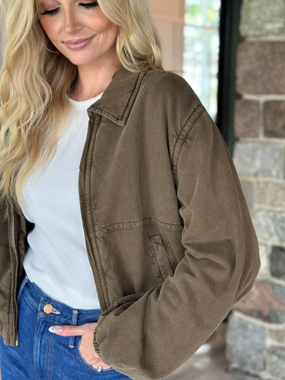 A woman wearing a brown corduroy jacket over a white ribbed top and blue jeans, standing in front of a stone wall.