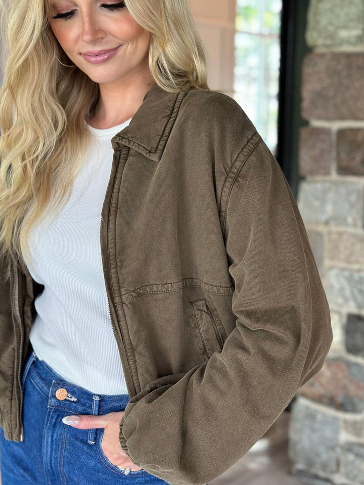A woman wearing a brown corduroy jacket over a white ribbed top and blue jeans, standing in front of a stone wall.