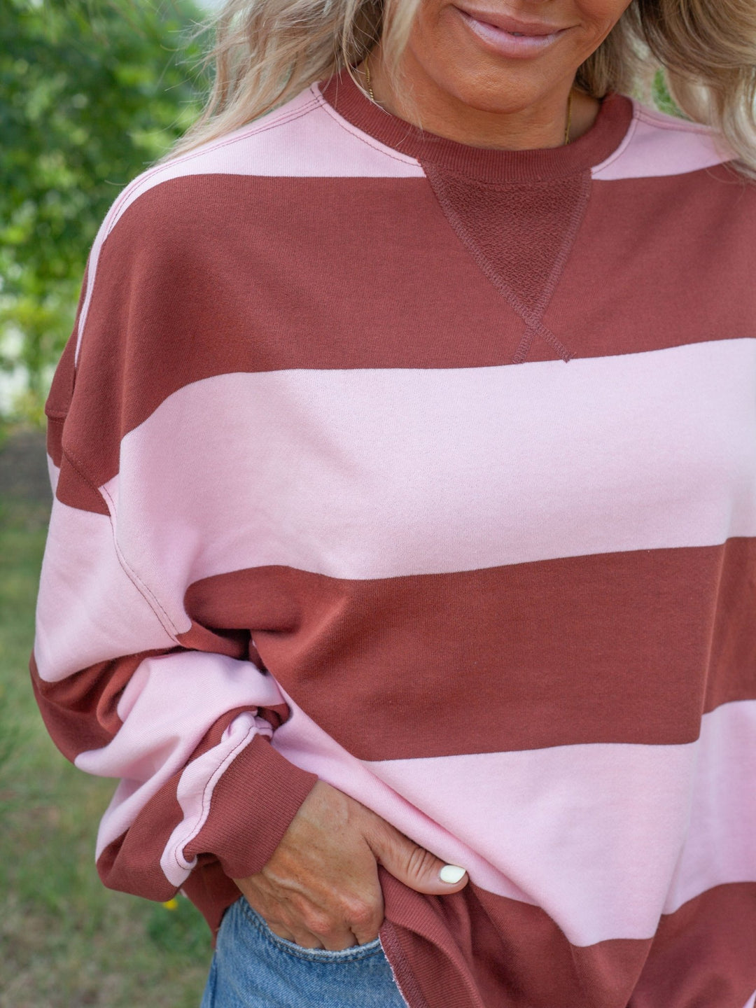 A close-up of a woman wearing a long-sleeve crewneck sweatshirt with wide horizontal stripes in dusty rose and light pink, paired with blue jeans.