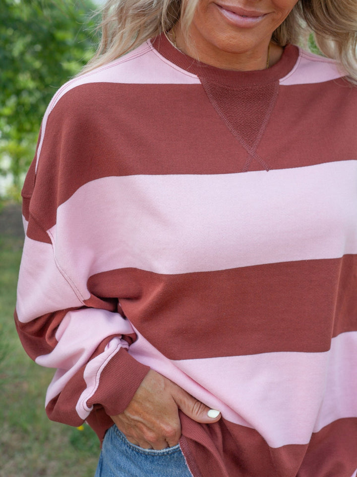 A close-up of a woman wearing a long-sleeve crewneck sweatshirt with wide horizontal stripes in dusty rose and light pink, paired with blue jeans.