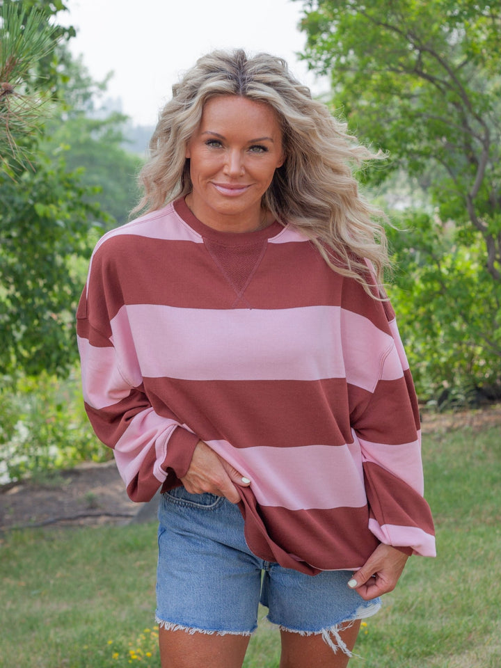 A woman wearing a long-sleeve crewneck sweatshirt with wide horizontal stripes in dusty rose and light pink, paired with denim shorts, standing outdoors in a grassy area with trees in the background.