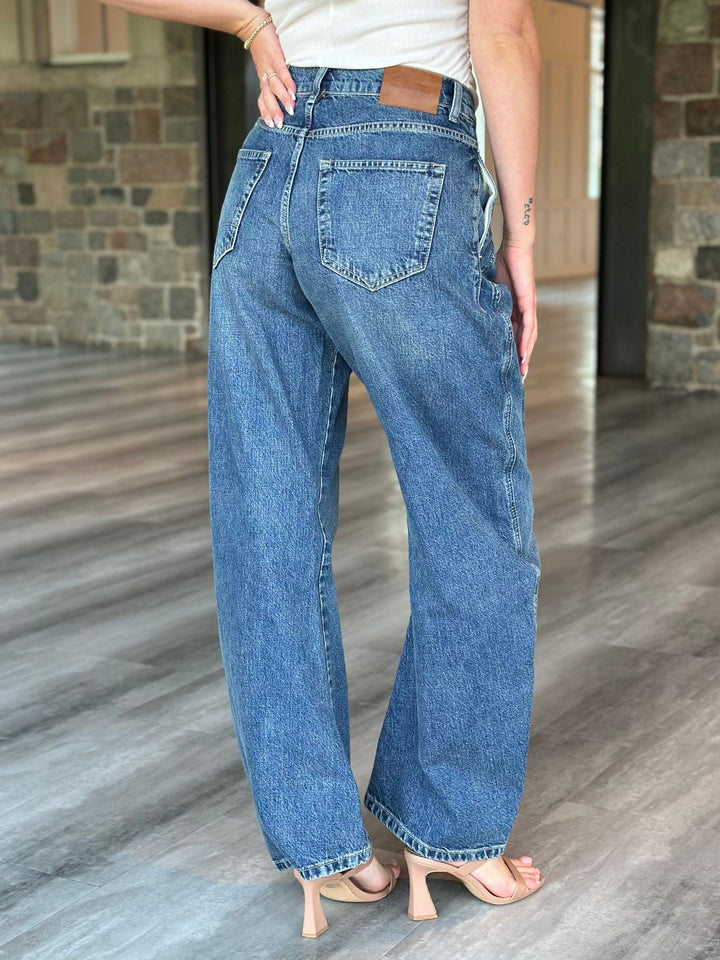 A person shown from the back wearing wide-leg blue denim jeans with a white top and nude heeled sandals, standing on a wood floor in front of a stone wall.