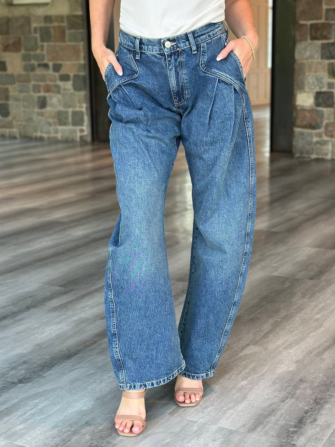 A person wearing a pair of wide-leg blue denim jeans with a relaxed, slightly baggy fit, standing on a gray wood-look floor in front of a stone wall.