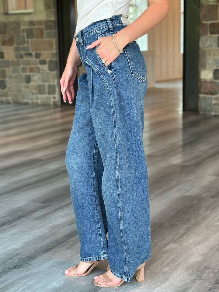 A person wearing high-waisted, wide-leg blue denim jeans with a hand in the pocket, standing on a gray wood floor in an indoor setting with stone walls.
