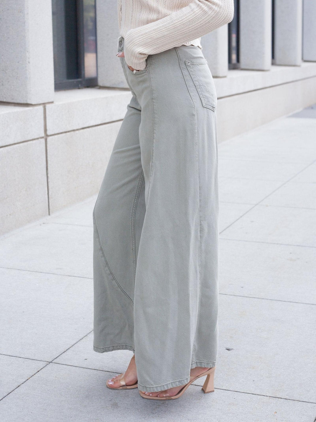 A person wearing light sage green wide-leg pants, a cream ribbed long-sleeve top, and clear heeled sandals, standing outdoors on a light concrete sidewalk.