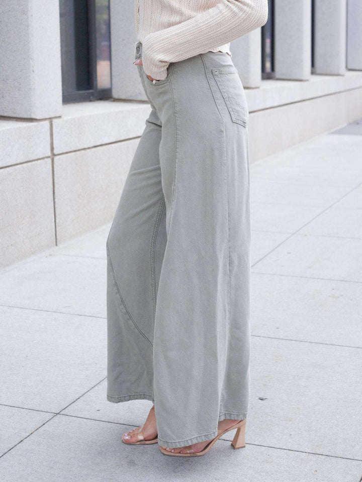 A person wearing light sage green wide-leg pants, a cream ribbed long-sleeve top, and clear heeled sandals, standing outdoors on a light concrete sidewalk.