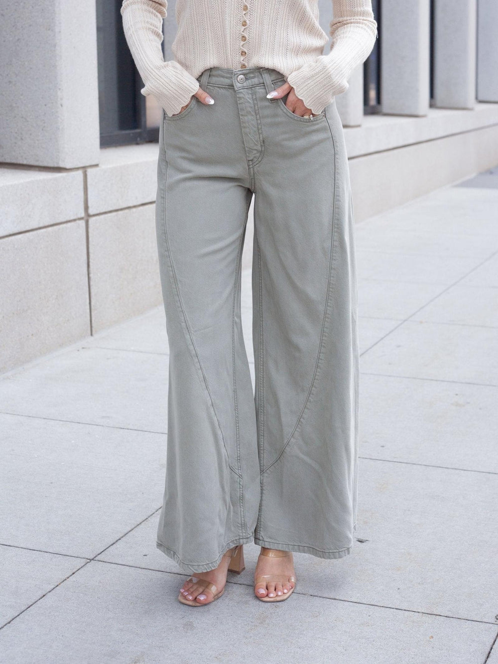 A person wearing light sage green wide-leg pants with a cream ribbed long-sleeve top and clear-strap heels, standing on a light concrete sidewalk.