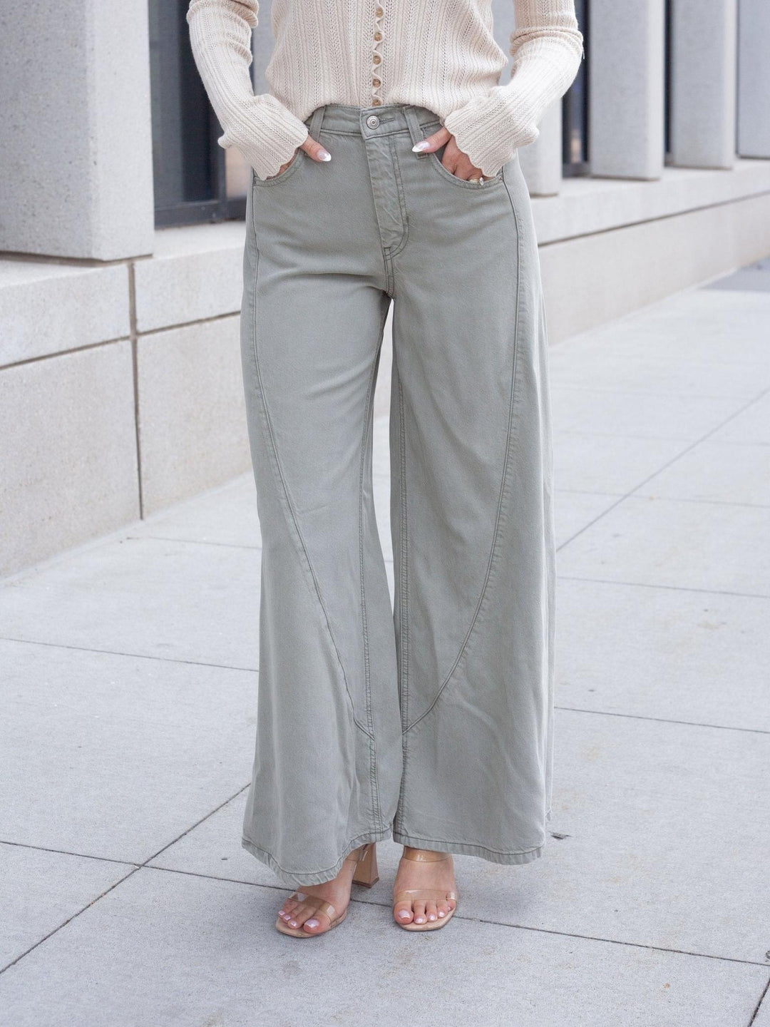 A person wearing light sage green wide-leg pants with a cream ribbed long-sleeve top and clear-strap heels, standing on a light concrete sidewalk.