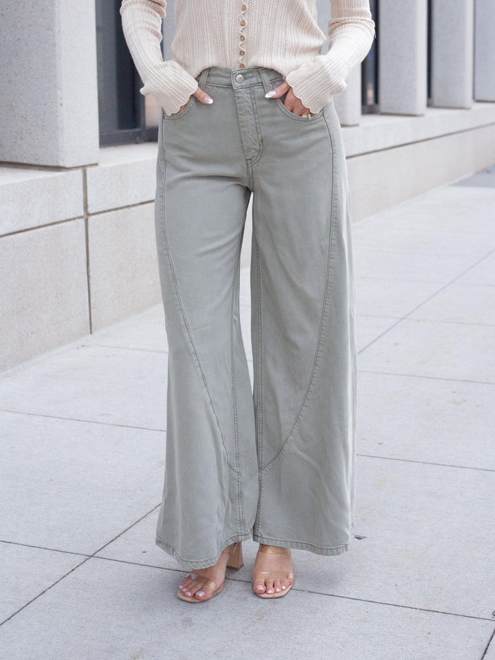 A person wearing light sage green wide-leg pants with a cream ribbed long-sleeve top and clear-strap heels, standing on a light concrete sidewalk.