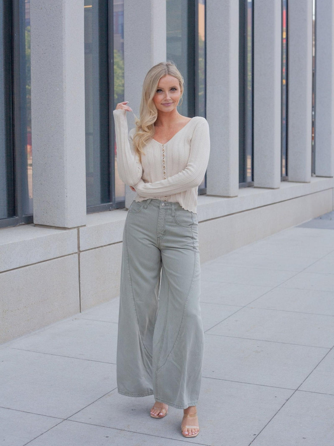 A woman stands outdoors in front of a modern building with large columns, wearing a cream long-sleeve knit top and light sage green wide-leg pants.