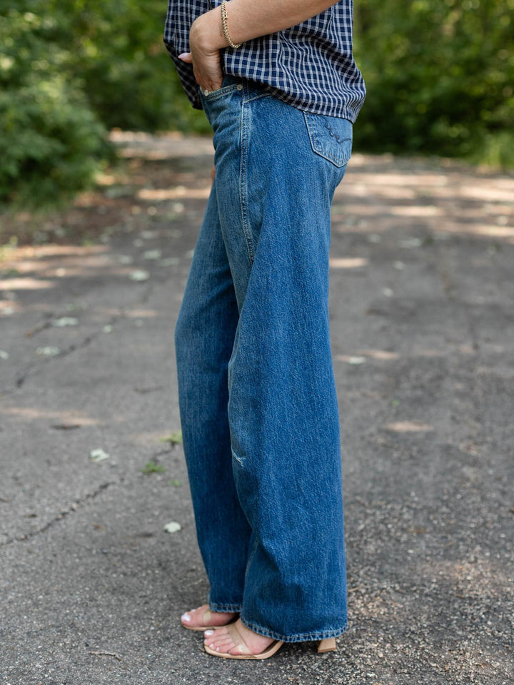A person wearing wide-leg blue denim jeans, a blue and white checkered shirt, and pink sandals, standing on an outdoor paved path with green foliage in the background.