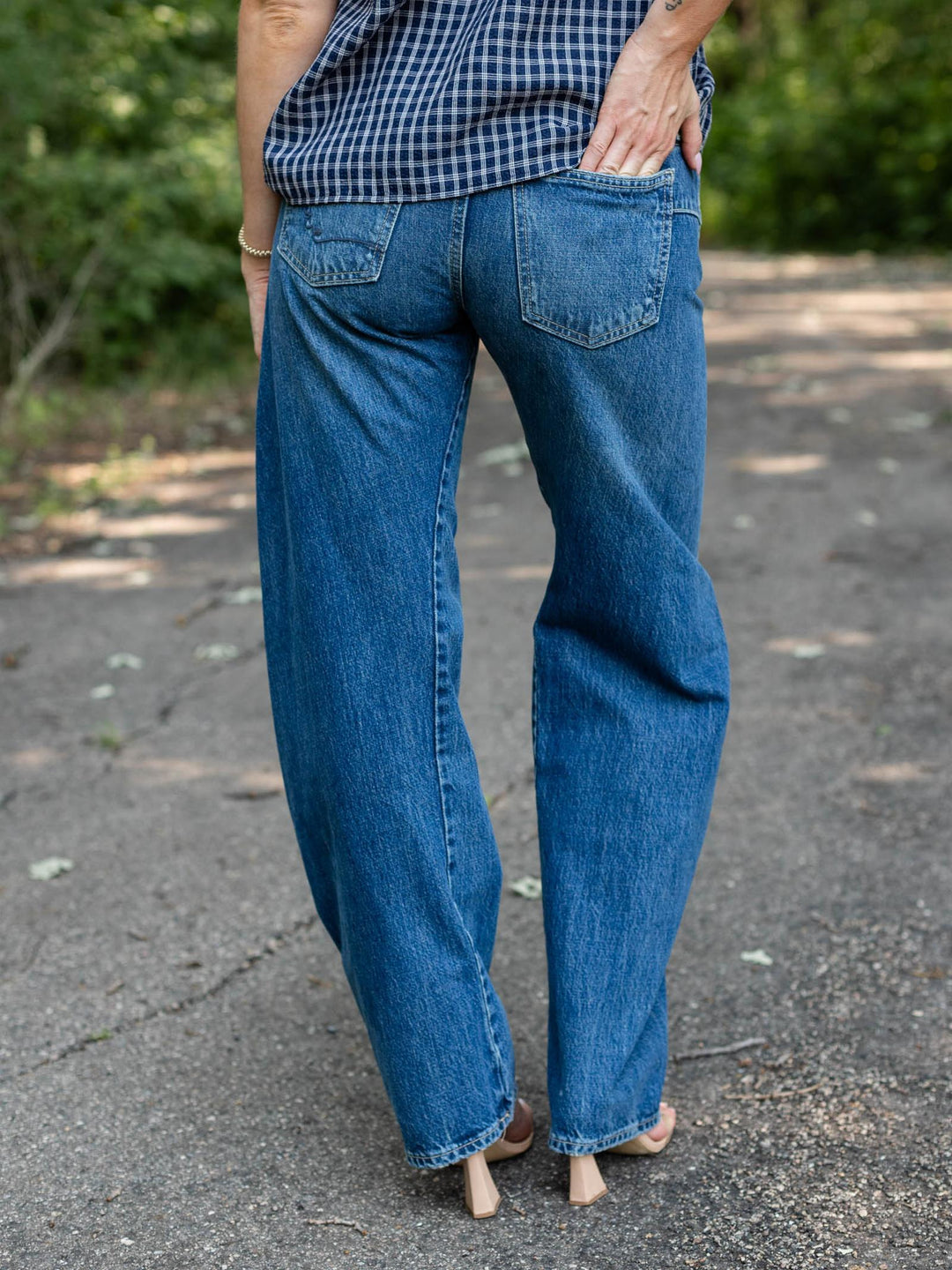 A person shown from the back wearing loose-fit blue denim jeans and a blue-and-white gingham top, standing outdoors on a paved path with blurred greenery in the background.