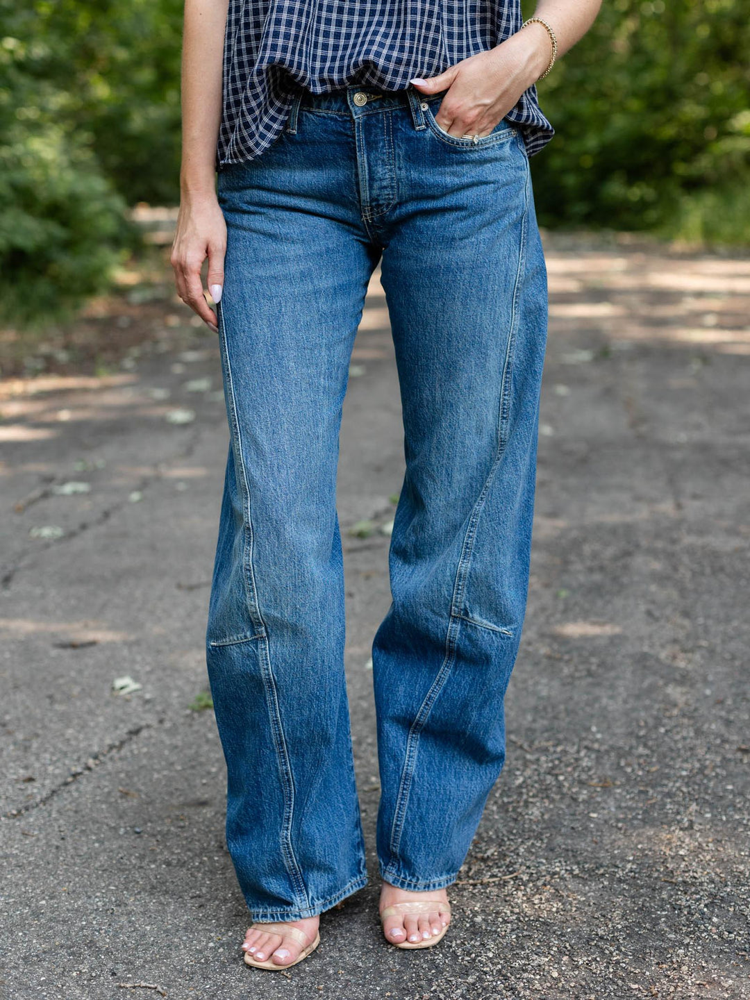 A person wearing medium-wash blue denim jeans with a relaxed, wide-leg fit, standing outdoors on a paved path with green foliage in the background.