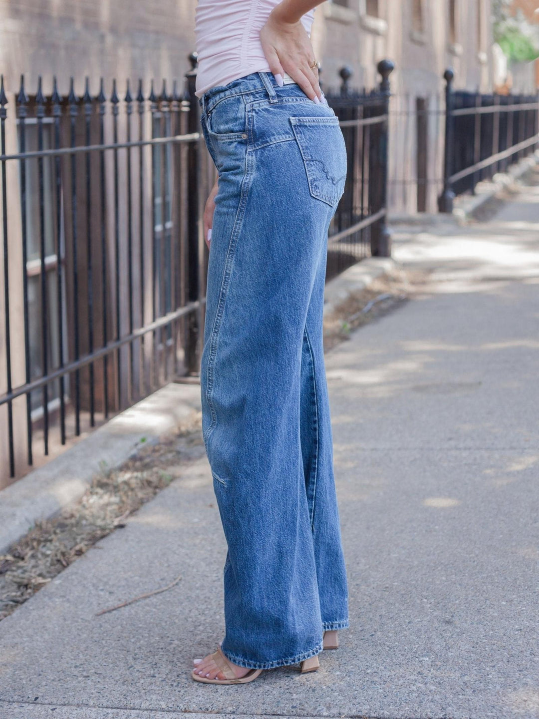 A person wearing high-waisted, wide-leg blue denim jeans with a light pink top, standing on a city sidewalk next to a black metal fence.