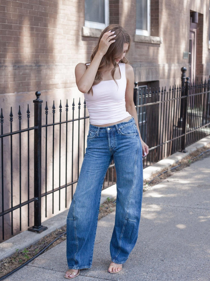 A woman stands on a sidewalk in front of a brick building and black metal fence, wearing a white ribbed tank top and high-waisted, wide-leg blue jeans.