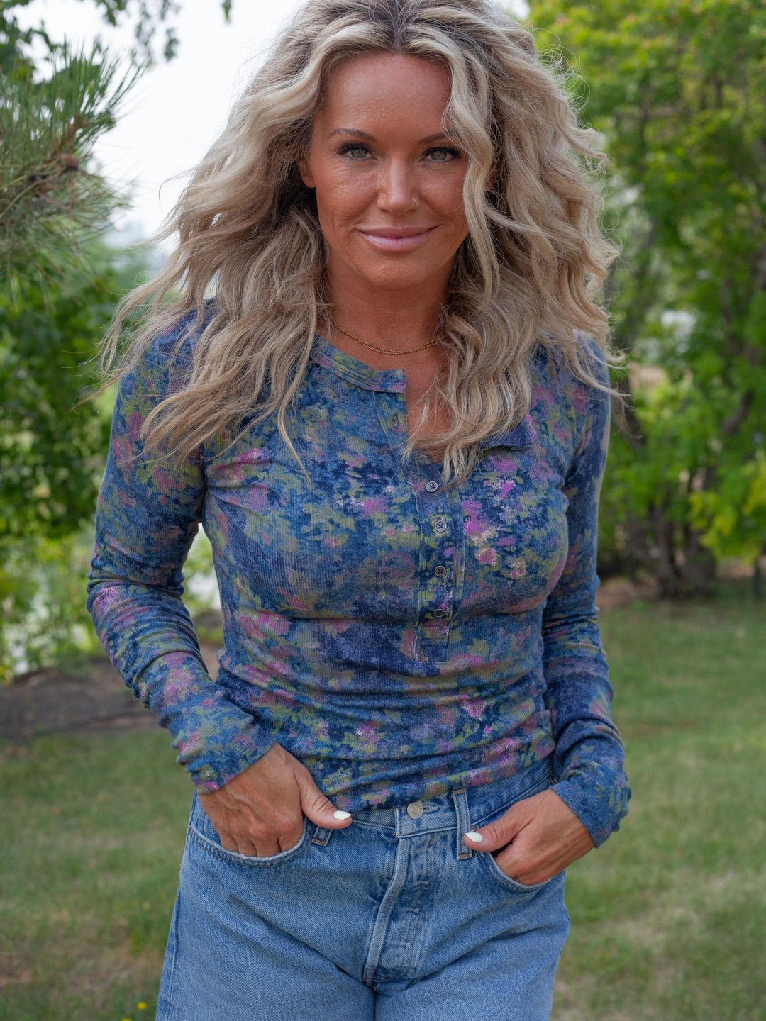A woman with long blonde hair stands outdoors wearing a long-sleeve henley top with a blue, purple, and pink floral pattern, paired with blue jeans.