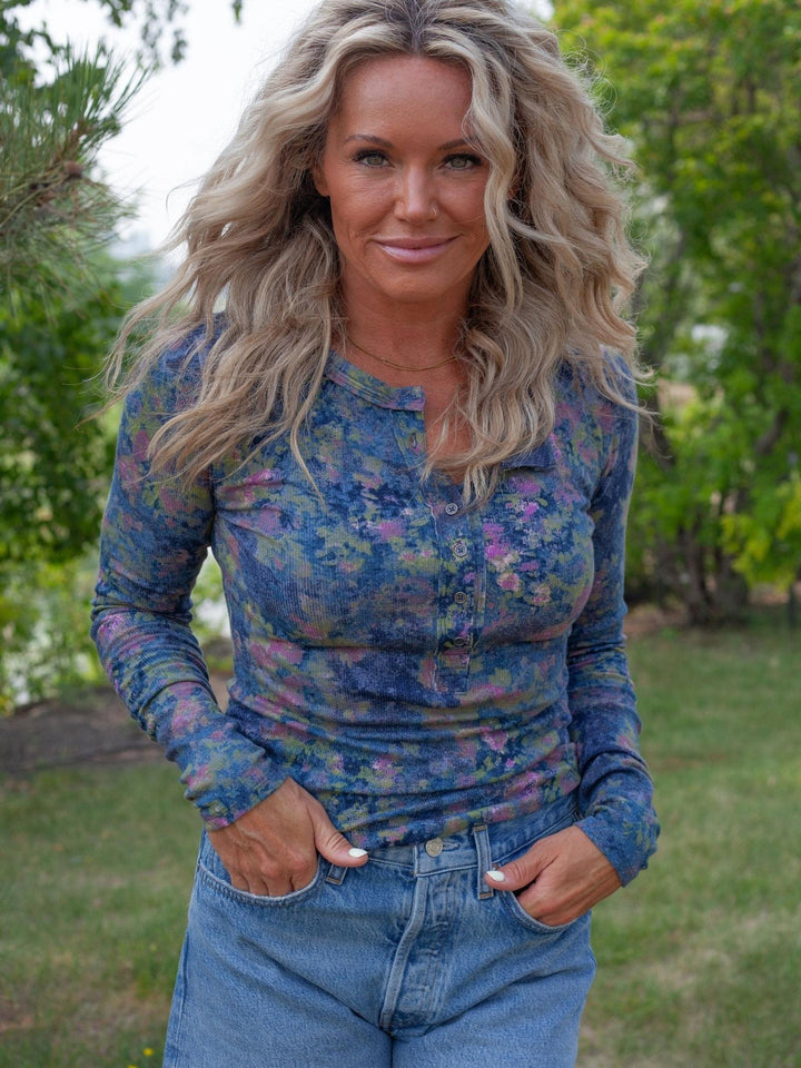 A woman with long blonde hair stands outdoors wearing a long-sleeve henley top with a blue, purple, and pink floral pattern, paired with blue jeans.