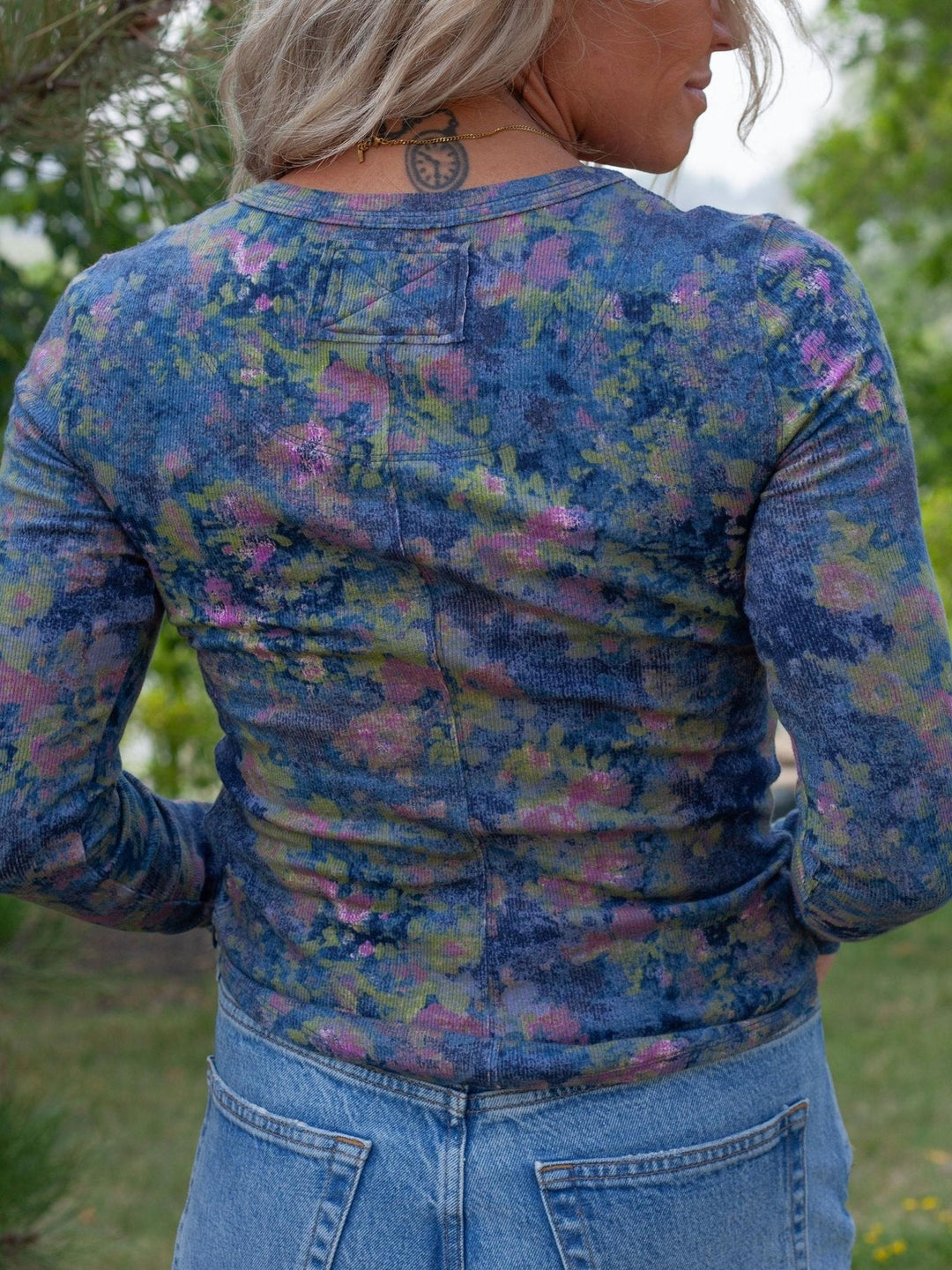 A woman viewed from behind wearing a fitted long-sleeve top with a blue floral print and light blue jeans, standing outdoors in a grassy area with trees in the background.