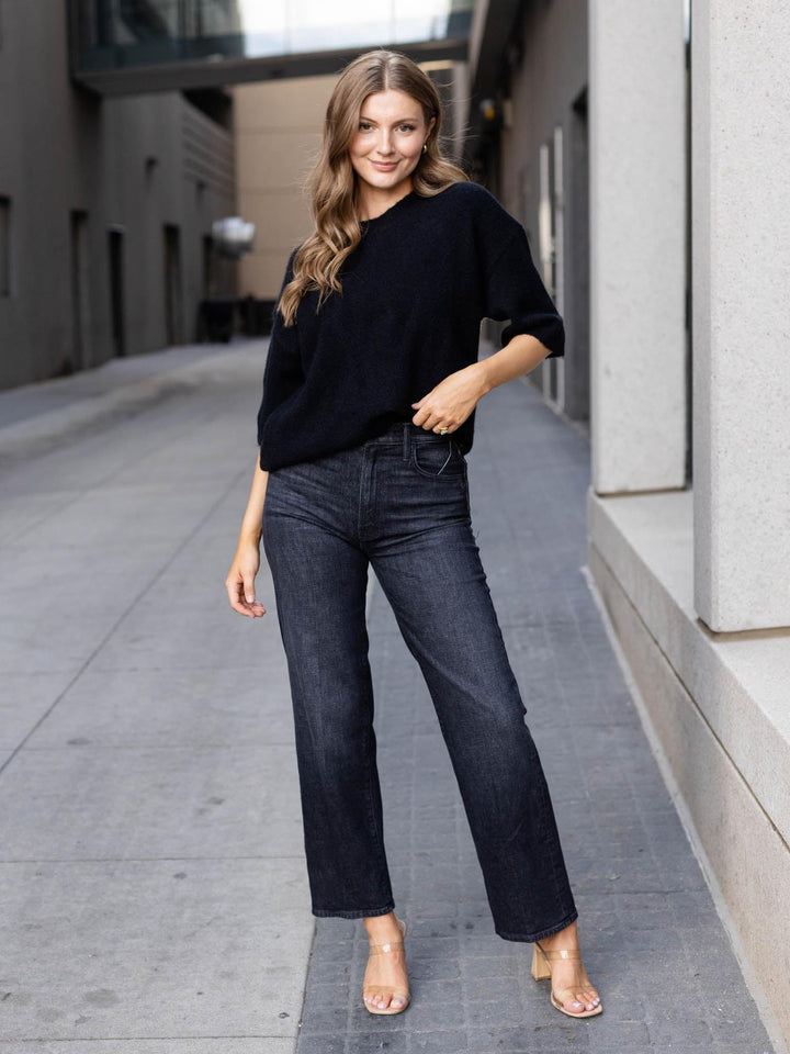 A woman stands on a city sidewalk wearing a black knit sweater, dark blue straight-leg jeans, and clear high-heeled sandals.