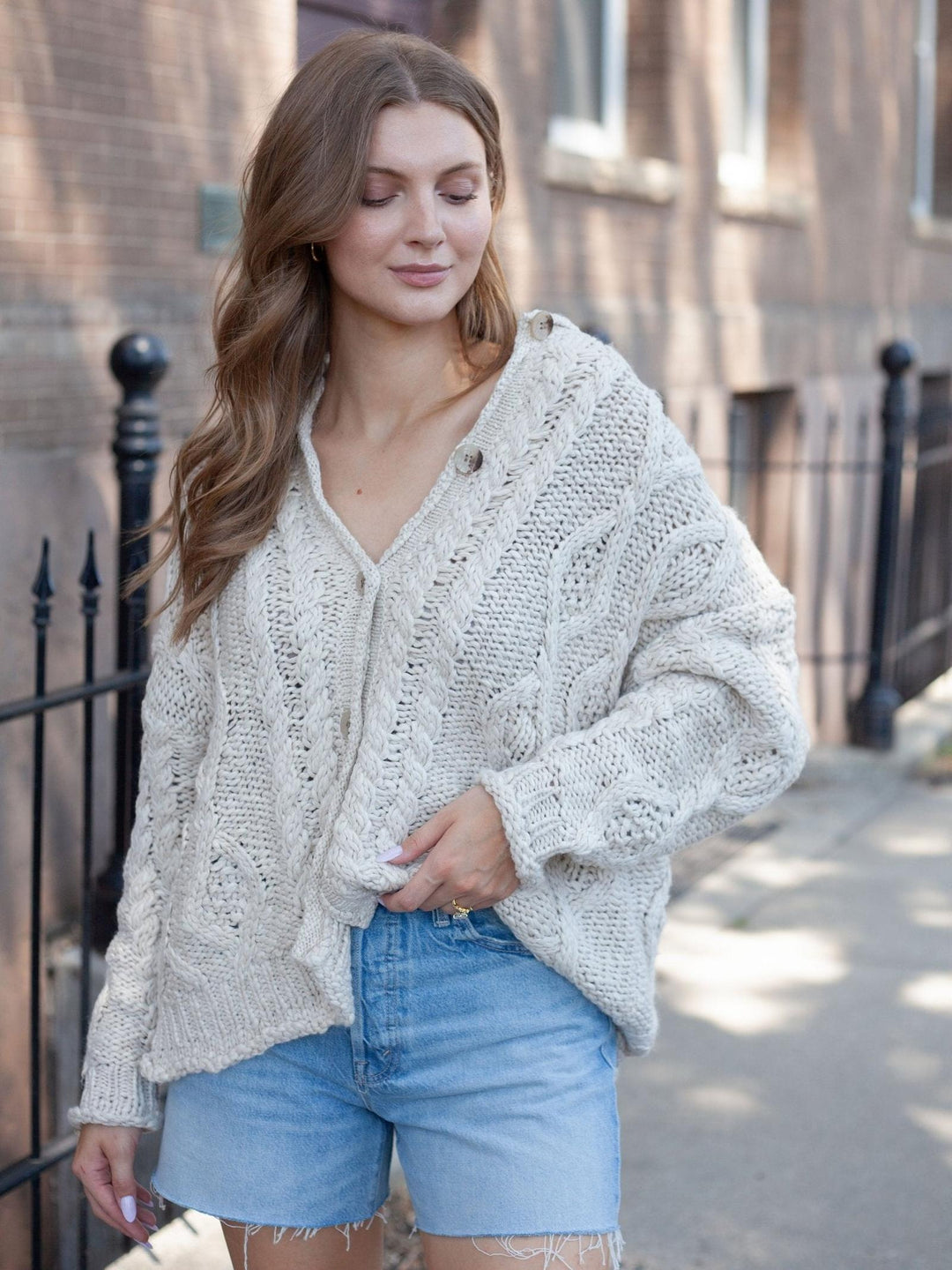 A woman wearing a chunky knit cream cardigan and light blue denim shorts, standing outdoors on a sidewalk in front of a brick building.