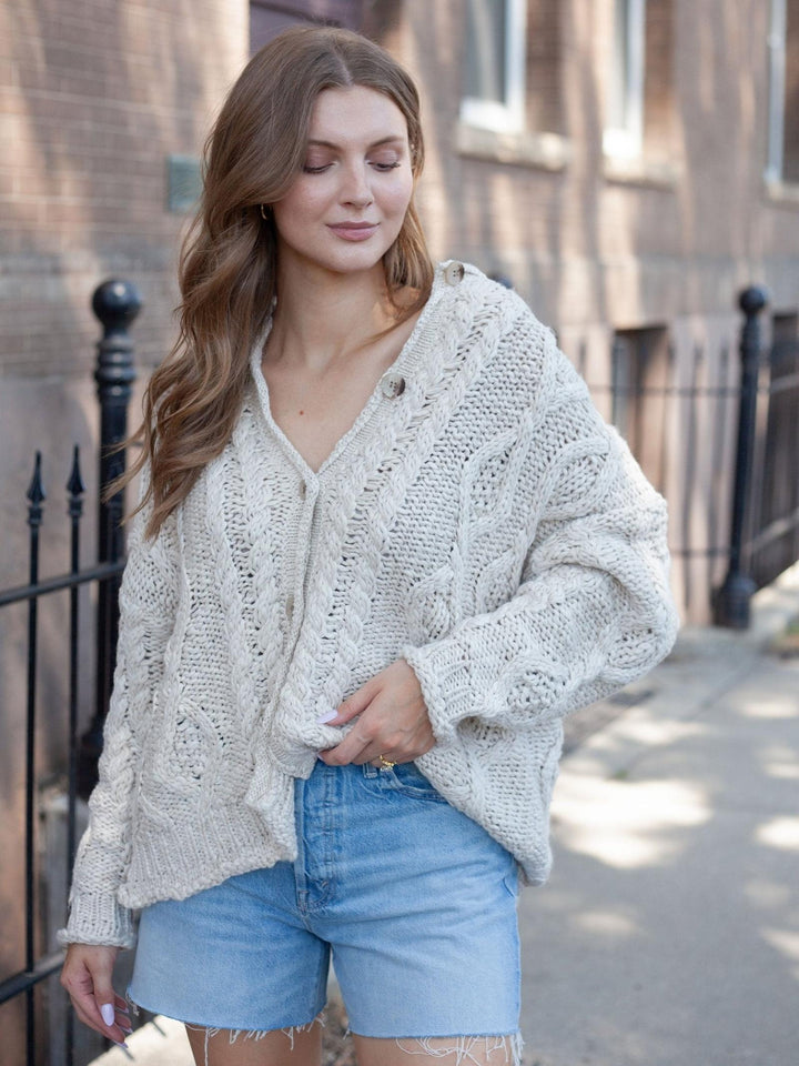 A woman wearing a chunky knit cream cardigan and light blue denim shorts, standing outdoors on a sidewalk in front of a brick building.