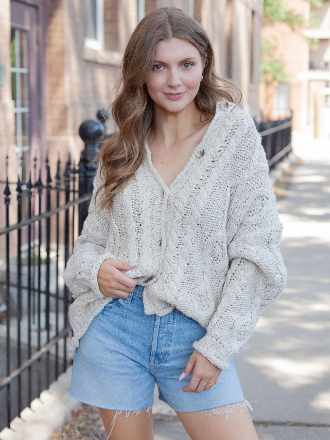 A woman wearing a chunky knit cardigan and denim shorts standing on an outdoor sidewalk in front of a brick building and black fence.