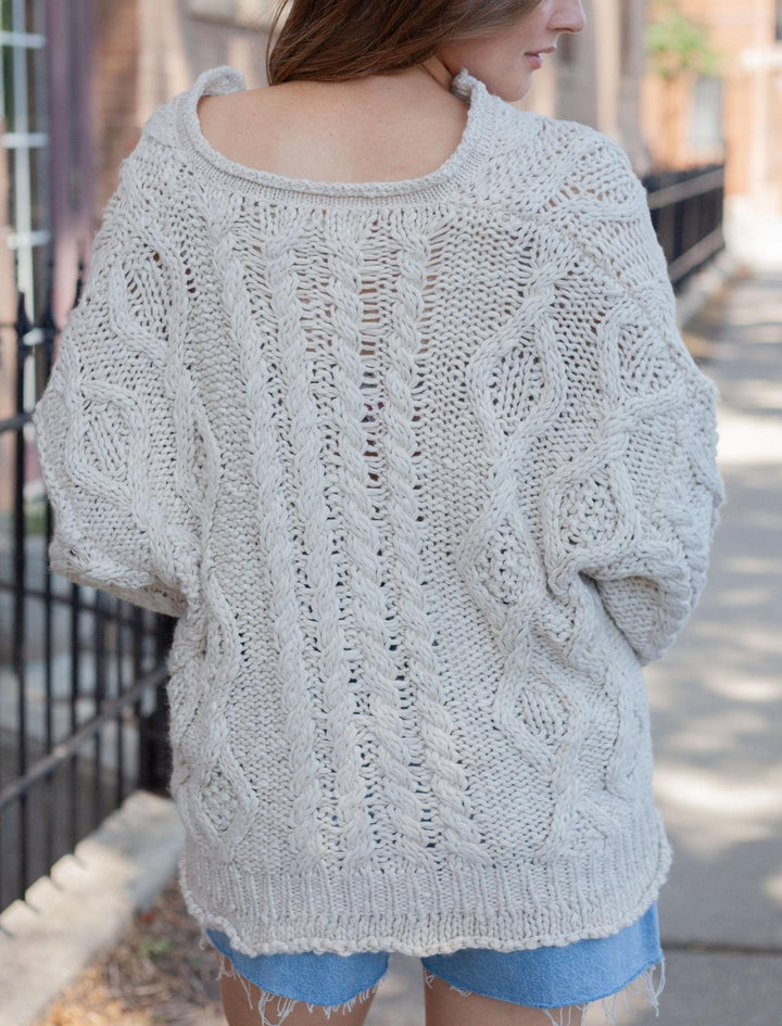 A woman wearing an oversized cream cable-knit sweater, shown from the back in an outdoor urban setting.