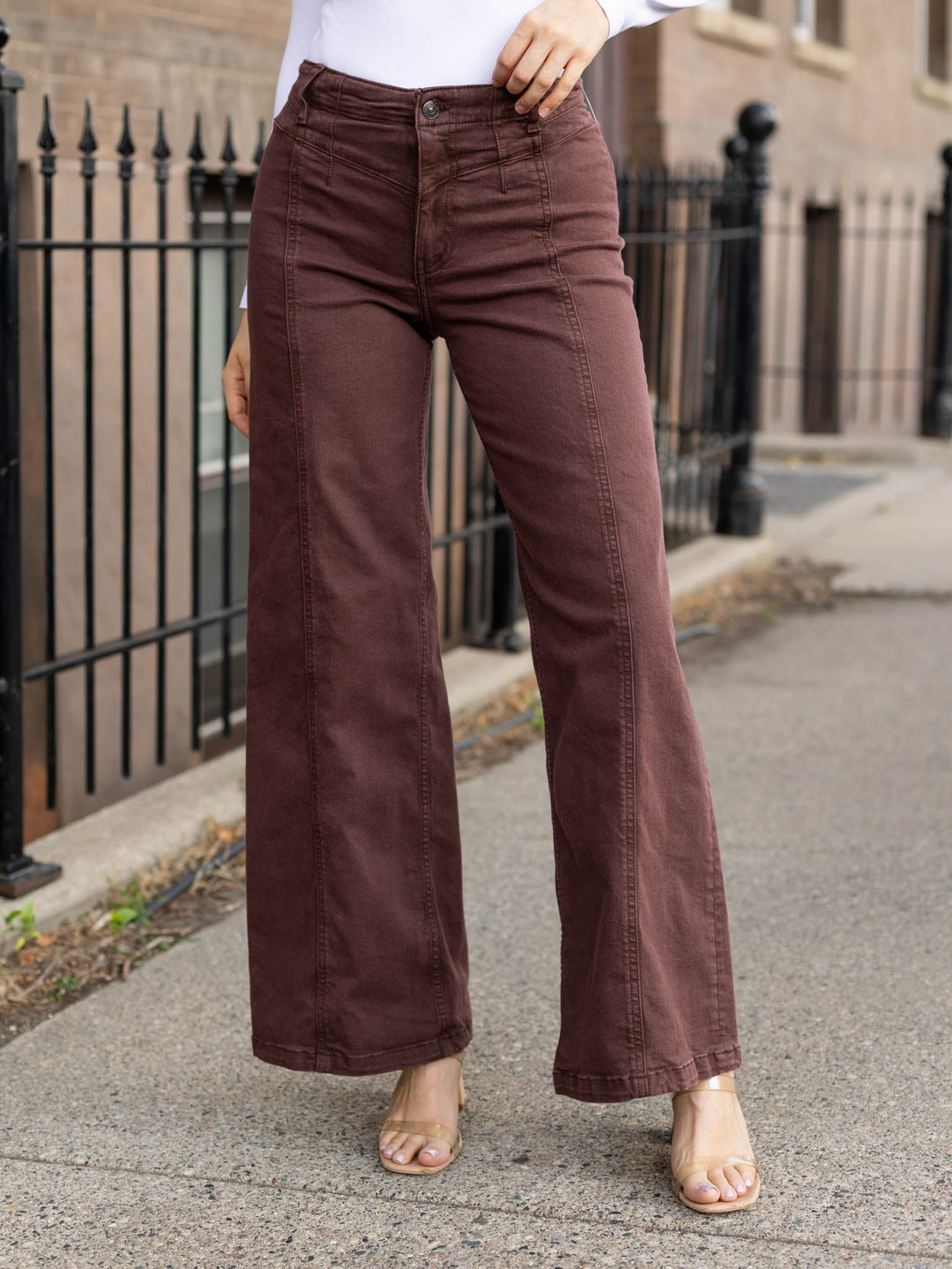 A person wearing high-waisted, wide-leg brown pants with a white top and clear high-heeled sandals, standing on a sidewalk in front of a brick building and black metal fence.