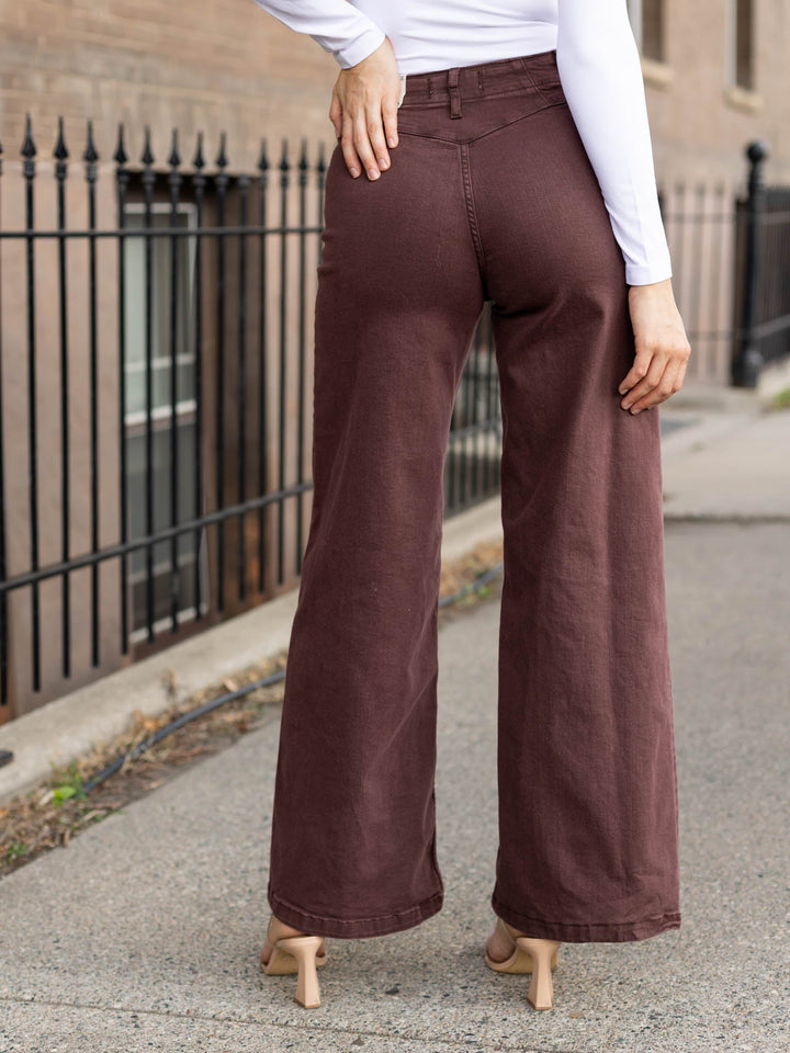 A person shown from the back wearing wide-leg brown pants, a white long-sleeve top, and beige heels on an outdoor sidewalk next to a black metal fence.
