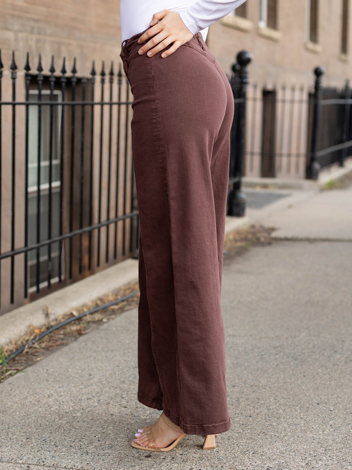 A person wearing high-waisted, wide-leg brown pants and clear high-heel sandals, standing on a city sidewalk next to a black metal fence.