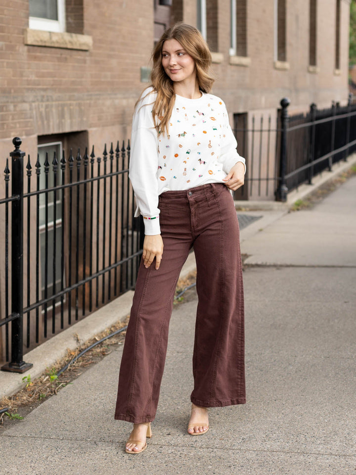A woman stands on a sidewalk in front of a brick building, wearing a white long-sleeve crewneck sweatshirt with a colorful bird pattern, brown wide-leg pants, and beige heeled sandals.