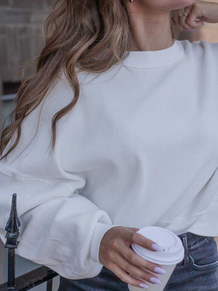 A woman wearing a white ribbed crewneck sweatshirt and dark gray jeans, holding a white paper coffee cup with a lid.
