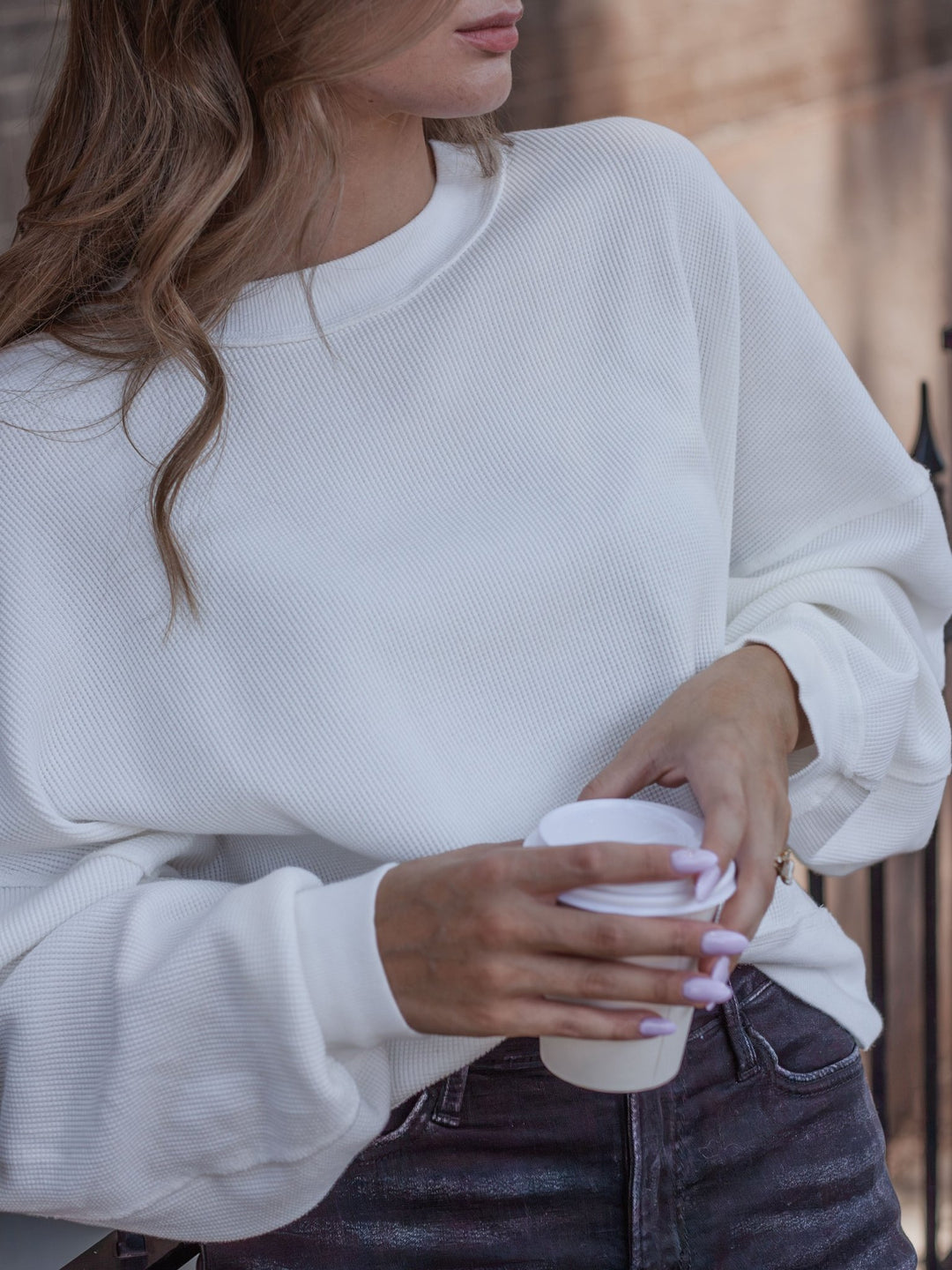 A woman wearing a white long-sleeve ribbed knit sweatshirt and dark gray jeans, holding a white paper coffee cup.