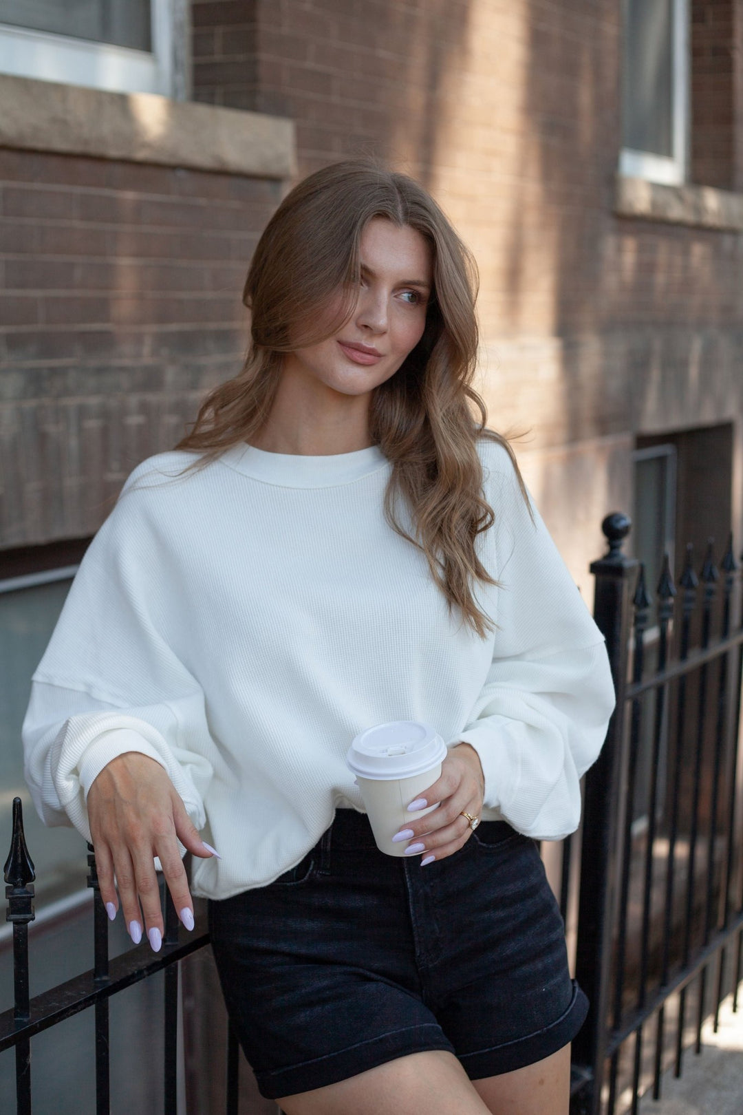 A woman with long wavy brown hair stands outdoors against a brick building, wearing a loose white crewneck sweatshirt and dark denim shorts while holding a white paper coffee cup.
