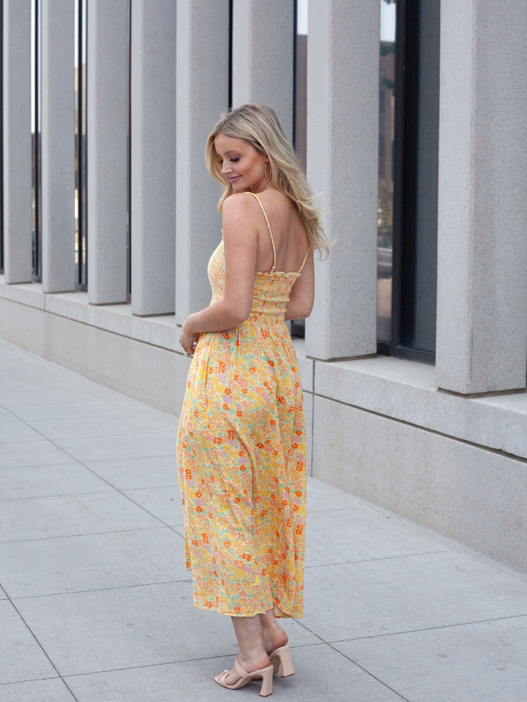 A woman wearing a yellow floral midi dress with spaghetti straps and a backless open back, standing on a paved walkway in front of a modern building with vertical columns.