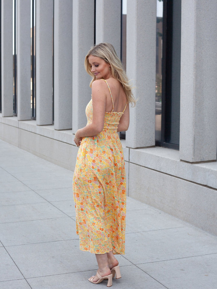 A woman wearing a yellow floral midi dress with spaghetti straps and a backless open back, standing on a paved walkway in front of a modern building with vertical columns.