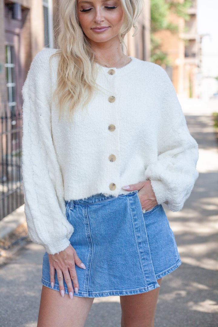A woman wearing a cream-colored, fuzzy long-sleeve button-up cardigan and a blue denim mini skirt, standing outdoors on a sidewalk.