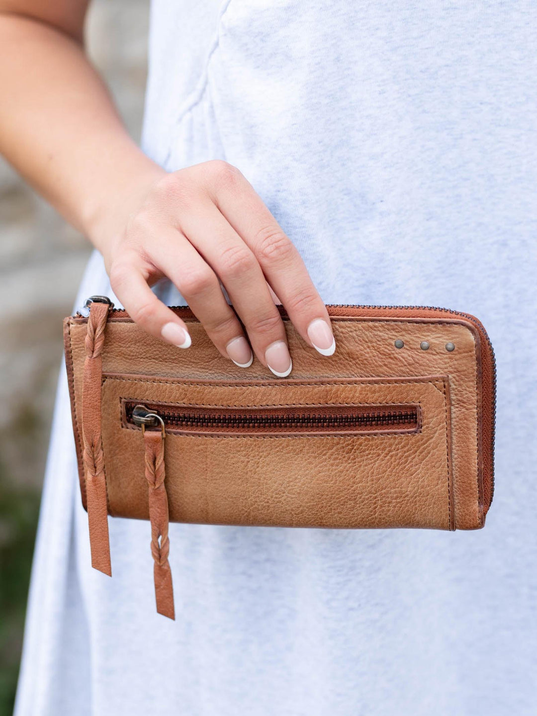 Hand holding a buttery-soft, warm Saddle Free People Emerson leather wallet with a front zip pocket and braided pull.