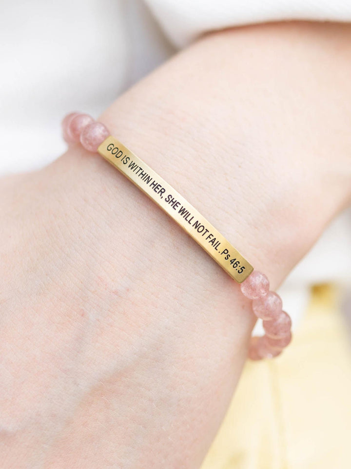 Pink Quartz stone bracelet with a gold bar engraved "GOD IS WITHIN HER, SHE WILL NOT FAIL. Ps 46:5" on a wrist.