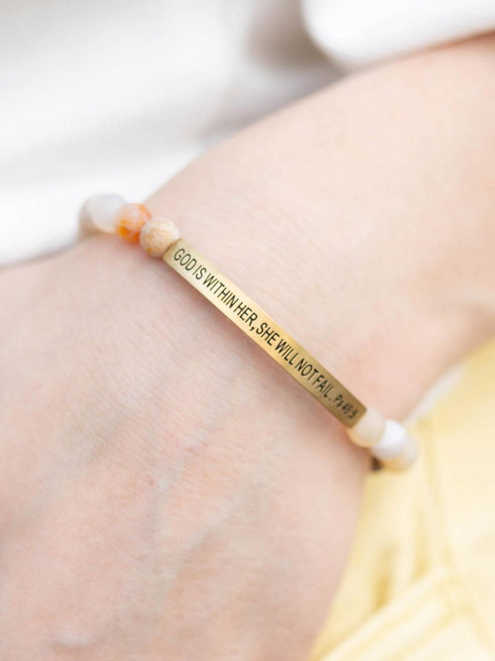 Close-up of a "God Is Within Her" stone bracelet with a gold bar and natural, earthy beads on a wrist.