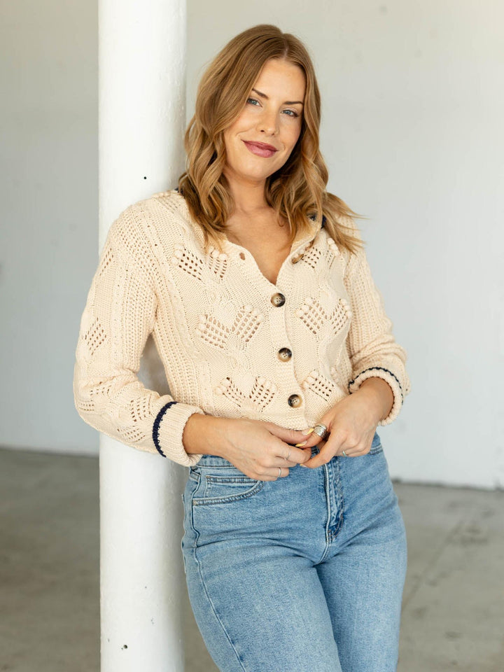 Woman wearing a beige knitted cardigan and blue jeans against a white wall.