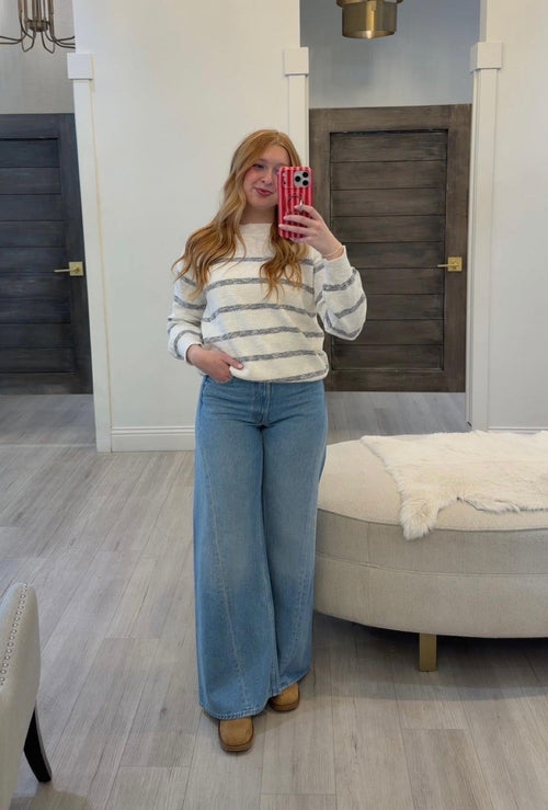 Woman taking a mirror selfie wearing a striped sweater and blue jeans in a room with wooden doors and a beige chair.