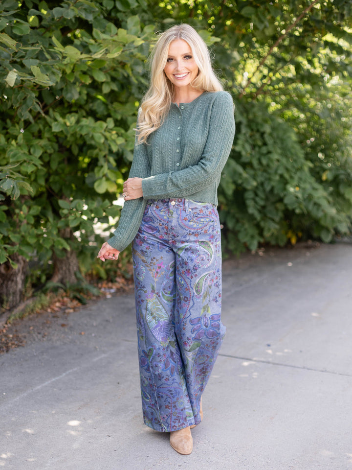 A woman standing outdoors on a paved path with green trees in the background, wearing a sage green knit cardigan and purple paisley wide-leg pants.