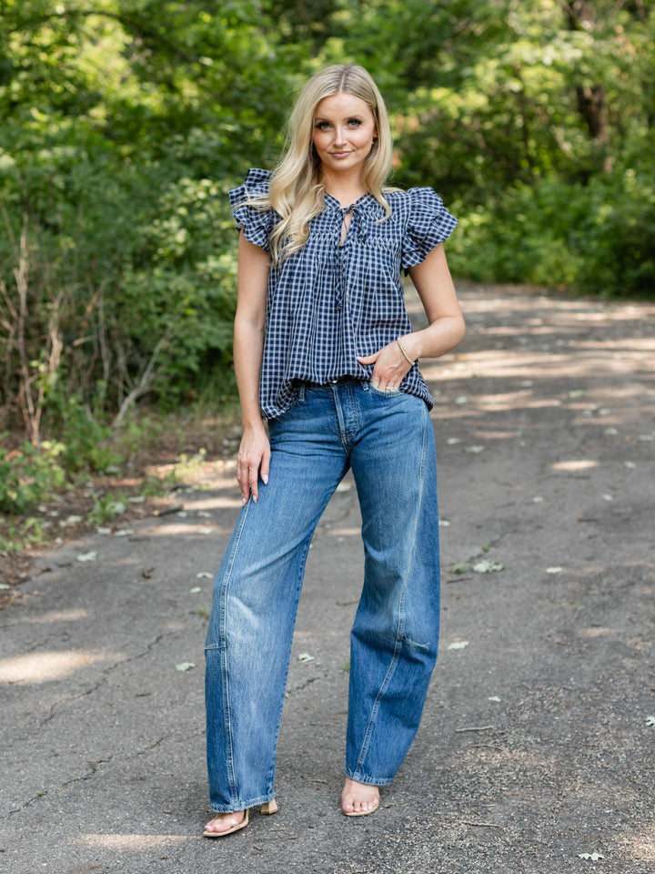 A woman stands on an outdoor paved path surrounded by green trees, wearing a blue and white gingham short-sleeve blouse with ruffled shoulders and blue wide-leg jeans.